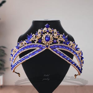 Puede incluir: Una tiara dorada con un diseño de doble banda, adornada con numerosas piedras preciosas azules y cristales transparentes. La tiara presenta una piedra azul ovalada central y se exhibe sobre una cabeza de maniquí negra. El texto "bridal jewelry" es visible.