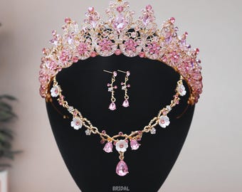 May include: A pink and gold bridal crown, necklace, and earrings set. The crown is adorned with pink and clear rhinestones and features a floral design. The necklace is a delicate chain with pink and white flowers and a teardrop pendant. The earrings are dangle earrings with pink and clear rhinestones.