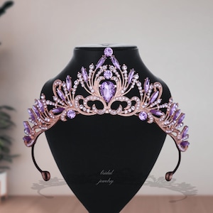 以下が含まれることがあります： 紫とクリアクリスタルが付いたローズゴールドのティアラ。ティアラの中央には大きな紫色のクリスタルがあり、小さな紫とクリアのクリスタルで飾られています。「bridal jewelry」の文字がディスプレイスタンドに書かれています。