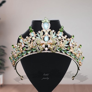 Può includere: Una tiara dorata ornata di pietre preziose verdi e trasparenti e accenti a goccia azzurri. La corona è esposta su una testa di manichino nera. Le parole "bridal jewelry" sono scritte in corsivo sul manichino.