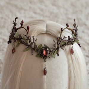 May include: A bronze-coloured crown with a celestial design. The crown features a central, large, rectangular, red gemstone, with a crescent moon on either side. The crown is adorned with branches, leaves, and dangling red teardrop-shaped gems.