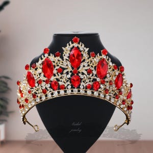 Op de afbeelding: Een goudkleurige kroon met grote rode, ovale edelstenen en kleinere rode en heldere kristallen. De kroon wordt getoond op een zwarte mannequinhoofd. De tekst "bridal jewelry" is zichtbaar.