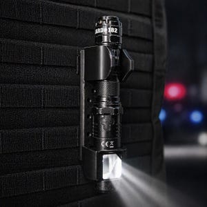 May include: A black tactical flashlight mounted on a black vest. The flashlight is cylindrical with a textured grip and a bright white light beam. The text "ARD #182" is visible on the flashlight.