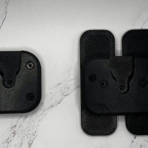 May include: Two black plastic mounting plates with screw holes and a raised lip around the edge. The plates are designed to be attached to a surface and have a slot in the centre for a mounting bracket.