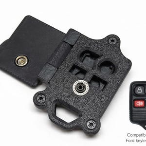 May include: Black key fob holder with a textured surface, open to reveal the interior. Includes a Ford keyless entry fob. The holder has a snap closure and is designed to hold a key fob.