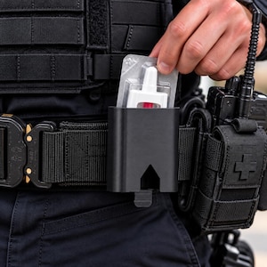 May include: A black tactical belt holster containing a white nasal spray in a clear blister pack. The holster is mounted on a black utility belt alongside a black radio pouch with a cross emblem and a black tactical vest with modular webbing.