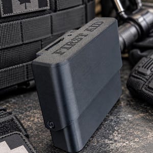 Puede incluir: Un estuche de primeros auxilios rectangular en negro mate con las palabras FIRST AID grabadas en la superficie superior. El estuche presenta una construcción rígida con un diseño articulado y un tornillo visible en el lateral, presentado en equipo táctico.