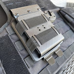 May include: A black tactical vest with several attached pouches and holders. The vest features straps, buckles, and elastic bands. The pouches are designed to hold equipment and accessories.