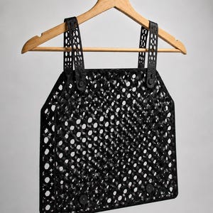 May include: Black, open-weave top with a geometric design, hanging on a wooden hanger. The top features a repeating pattern of small, circular holes and is held together with black straps and buttons. The garment is made of a flexible material.
