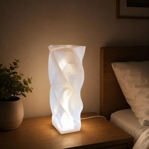 May include: A tall, translucent white table lamp with a spiraling, wave-like cylindrical structure. The lamp emits a warm, soft glow from within, highlighting the textured, layered surface of the material. It features a white power cord.