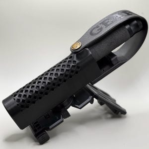 Könnte beinhalten: Schwarzes Pistolenholster mit einem schwarzen Lederriemen und einem goldenen Metallknopf. Der Riemen trägt den Aufdruck "GEAR". Das Holster hat ein schwarzes, rautenförmiges Netzmuster.