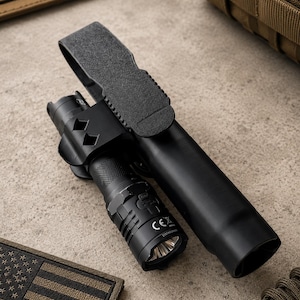 May include: A black tactical flashlight secured in a matching black polymer holster. The flashlight features a textured grip, a metallic reflector, and a CE marking printed on the side. The holster includes a wide belt clip attachment.