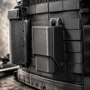May include: A black tactical vest featuring a grid of MOLLE webbing. Attached to the front are two black rigid polymer pouches. The pouch on the right has a hinged lid mechanism, while the pouch on the left has a textured, rectangular surface.