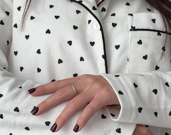 May include: A white long-sleeved button-down pajama top featuring a repeating pattern of small black hearts. The garment includes a notched collar with black piping trim along the edges and a single chest pocket on the left side.