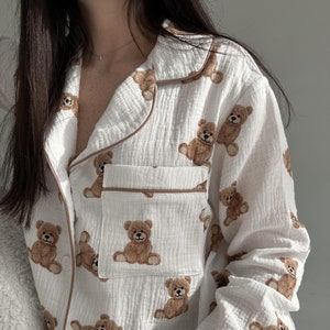Könnte beinhalten: Weißer Schlafanzug-Oberteil mit braunem Teddybär-Druck. Das Oberteil hat einen Knopfleisten-Verschluss und lange Ärmel.