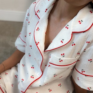 Può includere: Pigiama bianco con stampa a ciliegie rosse e bordatura rossa. La parte superiore ha un colletto abbottonato e maniche corte. I pantaloncini hanno una cintura elastica.