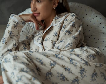 Puede incluir: Una mujer con una diadema blanca y un pijama de color crema con estampado floral azul. Está sentada en una cama con una almohada con estampado blanco y azul.