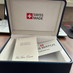 Puede incluir: Una caja de reloj rectangular de color azul marino con interior blanco. La tapa presenta un cuadrado rojo con una cruz blanca y el texto SWISS MADE. Dentro hay dos folletos blancos, etiquetados ETA G10.211 ETA G10.21A Eta Swiss Quartz y SWISS WATCH GUARANTEE.
