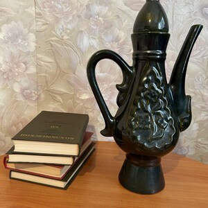 May include: A dark green ceramic teapot with a lid, handle, and spout. The teapot has a decorative relief pattern and sits on a wooden base. A stack of books is next to the teapot.