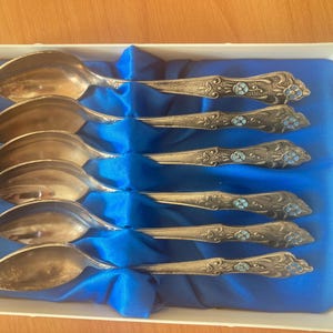 May include: Set of six silver teaspoons with blue enamel detailing. The spoons are arranged in a blue velvet box.