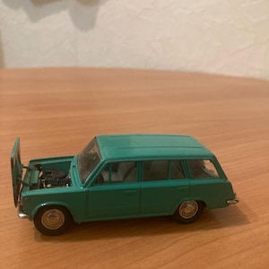 Puede incluir: Un modelo de coche de juguete azul verdoso con el capó abierto, que revela el motor. El coche tiene un estilo de carrocería familiar con una ventana trasera.