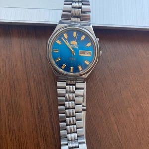 May include: A silver-toned Orient watch with a blue face and gold accents. The watch displays the day and date, with a silver band. The watch is on a wooden surface.