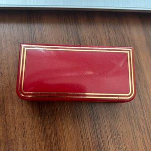 May include: A rectangular red jewelry box with a glossy finish and a double gold-toned border line detail on the lid. The box has rounded corners and a smooth, solid red surface, presenting a classic design for storing small accessories.