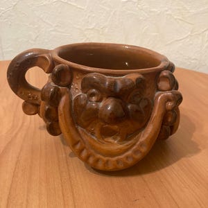 May include: A brown ceramic mug with a sculpted face and a handle. The mug has a textured surface and a wide rim.
