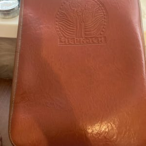 May include: A rectangular, brown leather case with a stitched border. The case features a circular embossed design with a figure and text in a foreign language. The leather has a slightly glossy finish.