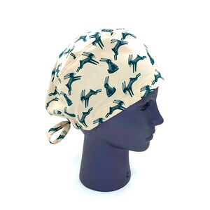 May include: A beige scrub hat with a green repeating pattern of stylized deer. The hat has a tie at the back.