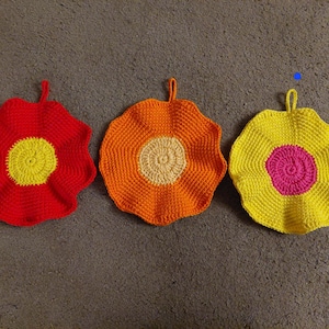 Könnte beinhalten: Drei handgefertigte Topflappen in Blumenform aus Häkelarbeit in leuchtenden Farben. Die Topflappen sind rot mit gelber Mitte, orange mit cremefarbener Mitte und gelb mit pinkfarbener Mitte. Jeder hat eine Schlaufe zum Aufhängen.