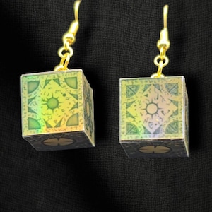 May include: A pair of gold-toned hook earrings featuring cube-shaped pendants with a green and black intricate geometric pattern. The text Hammer Family Jewels, Earrings, and Item BL2-45 is visible in blue font against the black background.