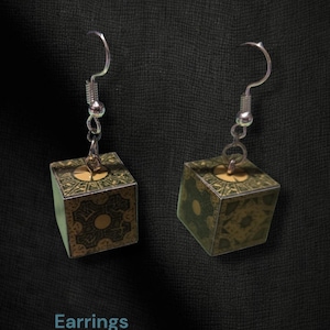 May include: A pair of dangle earrings featuring cube-shaped charms with intricate green and gold geometric patterns. The earrings hang from silver-toned metal hooks. The text Hammer Family Jewels, Earrings, and Item BL2-45 is visible in the image.