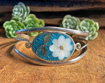 May include: Silver cuff bracelet with an oval turquoise inlay featuring a white flower design. The flower is made of mother-of-pearl. The bracelet has a twisted wire design and floral accents.