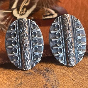 May include: A pair of oval-shaped silver earrings with intricate floral and geometric designs. The earrings have a dark, oxidized finish, highlighting the detailed patterns. The design includes vertical bands and decorative elements, creating a vintage aesthetic.
