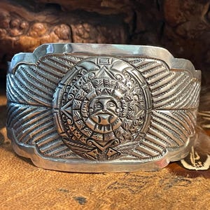 May include: A wide, silver-toned cuff bracelet with an intricate Aztec sun god design. The central medallion features a detailed face surrounded by radiating patterns and geometric shapes. The bracelet is approximately 2 inches wide.