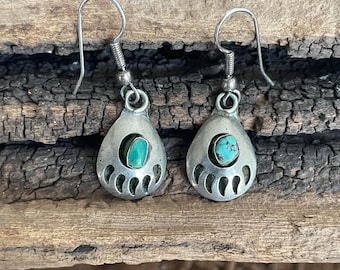 Pode incluir: Um par de brincos prateados com formato de gota. Cada brinco apresenta uma pedra turquesa no centro e um design de pata de urso ao redor da pedra. Os brincos têm um fecho de gancho.