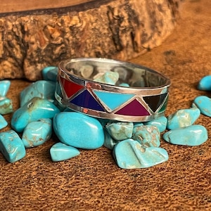May include: A silver ring with a geometric design featuring inlaid turquoise, red, purple, blue, and black stones.