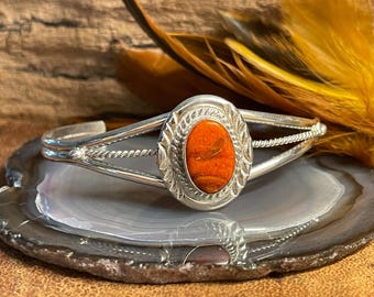 Pode incluir: Uma pulseira de prata com uma grande pedra oval laranja engastada em um bezel de prata. A pulseira tem um design de fio trançado e é aberta de um lado.