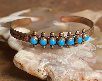 Pode incluir: Um bracelete de cobre com pedras turquesa e um design de flecha. O bracelete está sobre uma rocha branca e marrom.