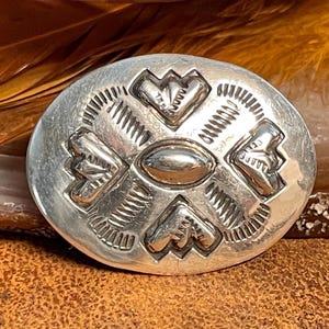 May include: A silver oval-shaped pendant with a geometric design featuring four stylized hearts and a central oval. The pendant has a textured surface and a slightly raised center.