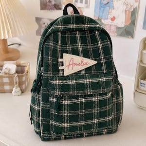 Op de afbeelding: Een donkergroene rugzak met een wit en zwart ruitpatroon. De tas is voorzien van een handvat aan de bovenkant, een voorvak met ritssluiting en een driehoekige crèmekleurige patch op de voorzijde met de naam Amelia in roze cursief geborduurd.