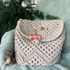 以下が含まれることがあります： ベージュのマクラメバックパック、フラップ開閉式。バッグは織り模様で、羽と金色の蜂の装飾チャームが付いています。調節可能なストラップは、白のストライプが入ったベージュです。