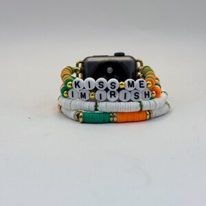 May include: Stack of bracelets with white, green, and orange beads, with the words "KISS ME I'M IRISH" spelled out in white letter beads. The bracelets are stacked around a black smartwatch.