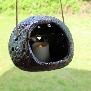 K&ouml;nnte beinhalten: Ein h&auml;ngender Kerzenhalter aus Keramik mit einer rauen, dunkelbraunen Textur und einem quadratischen Ausschnitt. Eine wei&szlig;e Kerze ist in den Ausschnitt gestellt und die Kerze brennt.