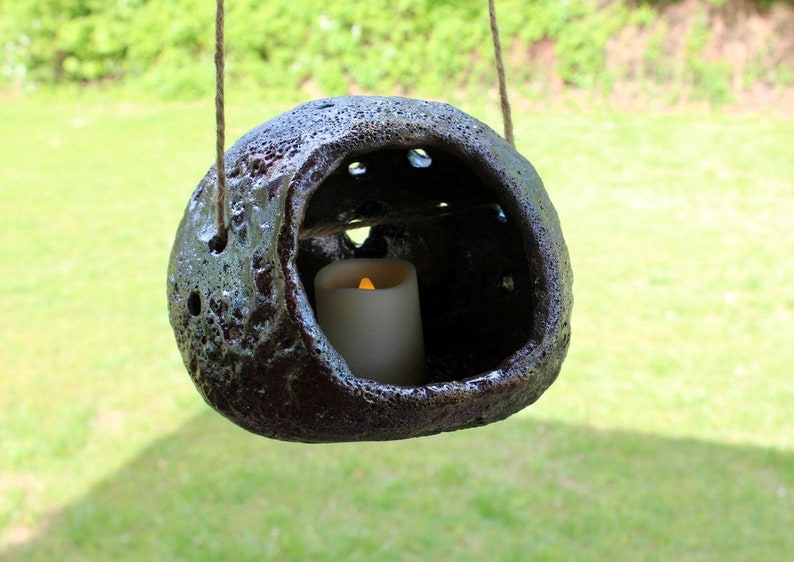 K&ouml;nnte beinhalten: Ein h&auml;ngender Kerzenhalter aus Keramik mit einer rauen, dunkelbraunen Textur und einem quadratischen Ausschnitt. Eine wei&szlig;e Kerze ist in den Ausschnitt gestellt und die Kerze brennt.