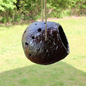 K&ouml;nnte beinhalten: Ein dunkelbraunes Vogelhaus aus Keramik mit einem Loch zum Betreten und einer Schnur zum Aufh&auml;ngen. Das Vogelhaus hat eine strukturierte Oberfl&auml;che und ist mit kleinen L&ouml;chern verziert.