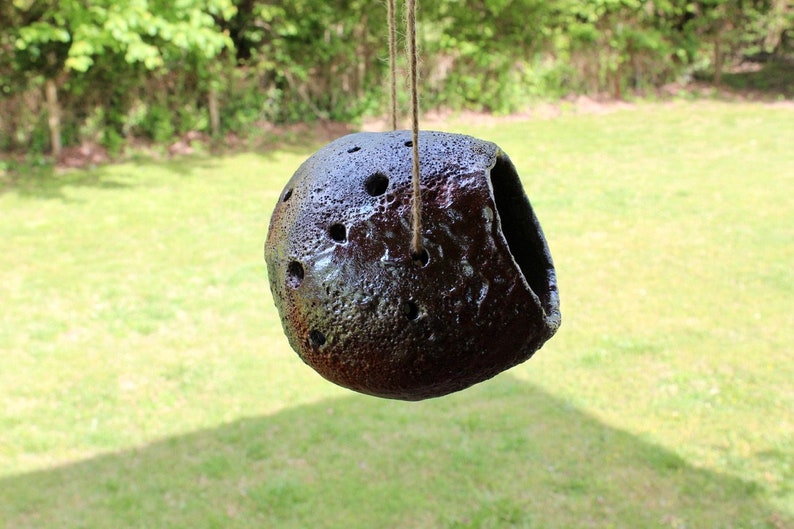 K&ouml;nnte beinhalten: Ein dunkelbraunes Vogelhaus aus Keramik mit einem Loch zum Betreten und einer Schnur zum Aufh&auml;ngen. Das Vogelhaus hat eine strukturierte Oberfl&auml;che und ist mit kleinen L&ouml;chern verziert.