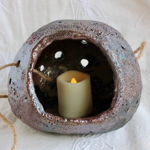 K&ouml;nnte beinhalten: Ein h&auml;ngender Kerzenhalter aus Keramik mit einer rauen, strukturierten Oberfl&auml;che und einer kleinen &Ouml;ffnung f&uuml;r eine Kerze. Der Halter ist aus braunem und grauem Ton gefertigt und hat eine Schlaufe aus Seil zum Aufh&auml;ngen. Eine kleine wei&szlig;e Kerze befindet sich im Halter.