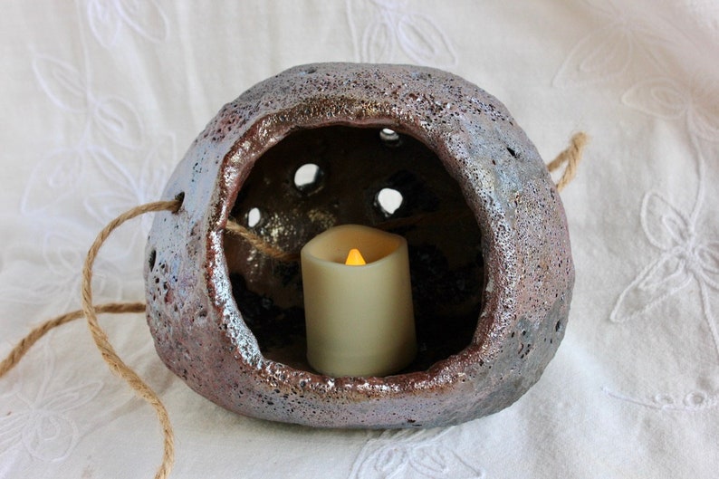 K&ouml;nnte beinhalten: Ein h&auml;ngender Kerzenhalter aus Keramik mit einer rauen, strukturierten Oberfl&auml;che und einer kleinen &Ouml;ffnung f&uuml;r eine Kerze. Der Halter ist aus braunem und grauem Ton gefertigt und hat eine Schlaufe aus Seil zum Aufh&auml;ngen. Eine kleine wei&szlig;e Kerze befindet sich im Halter.